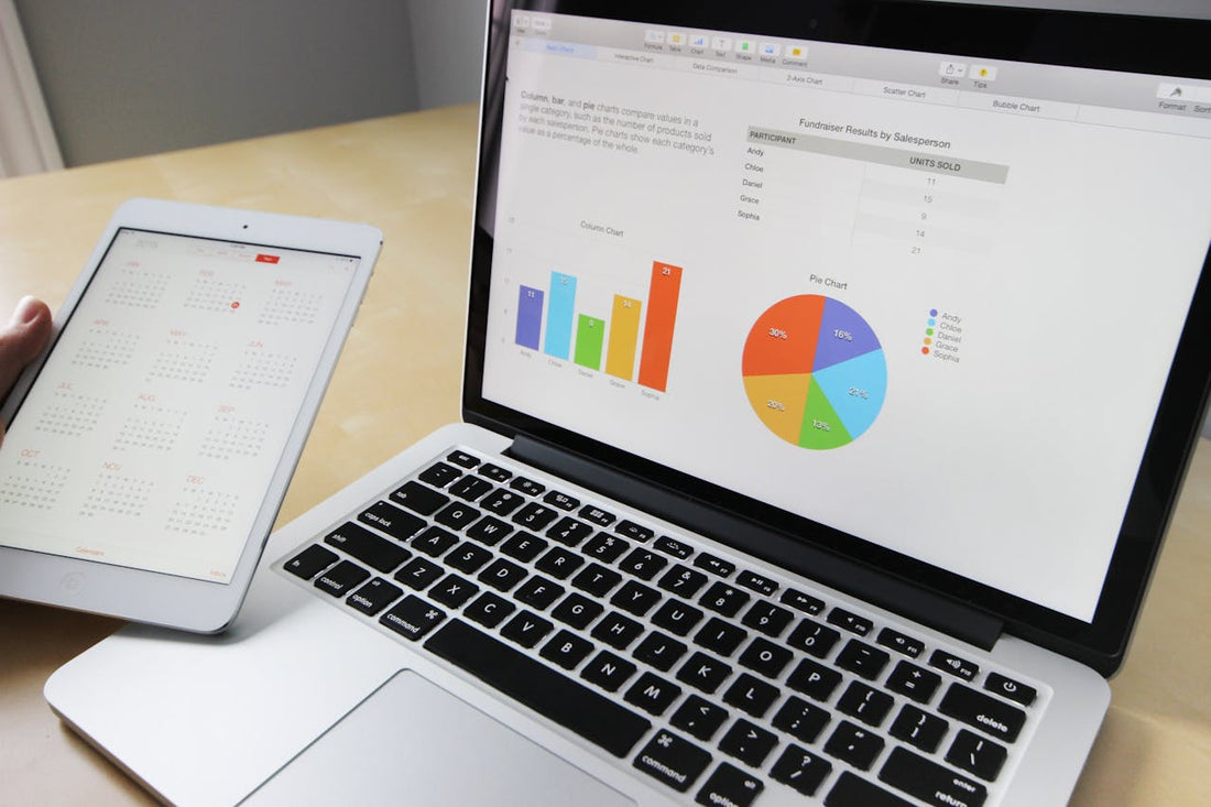 Doigt d’un individu sur une table tenant une tablette où est affiché un calendrier. Sur la table, se trouve un ordinateur avec des données analytiques (diagramme, camembert, tableau). 