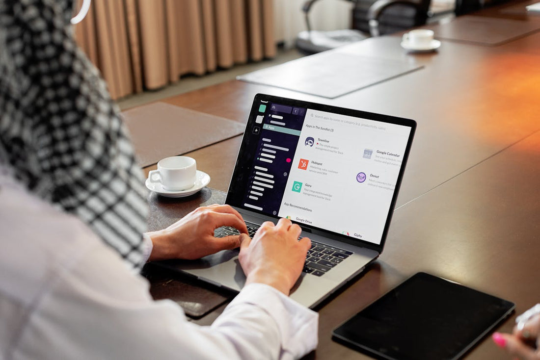 Individu dans un bureau les mains sur le clavier de son ordinateur. À ses côtés une tasse de café blanche, et une tablette noire. 