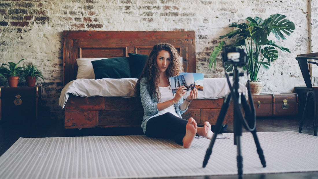 Femme assise sur le sol. Elle présente des photos à la caméra qui se tient sur un trépied noir. 